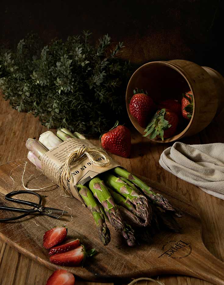 A bundle of asparagus sits on a wooden table with a bowl of strawberries tipped over, next to it.