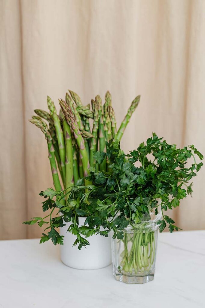 Two containers sit on a white tabletop. One holds fresh herbs, the other holds fresh asparagus.