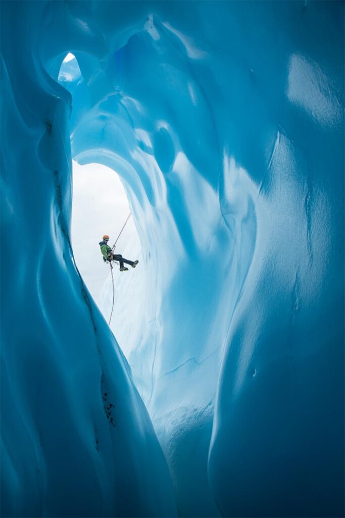 An ice climber in a green jacket and orange helmet rappels past a large rounded entrance to an ice cave on the Matanuska Glacier in Alaska.