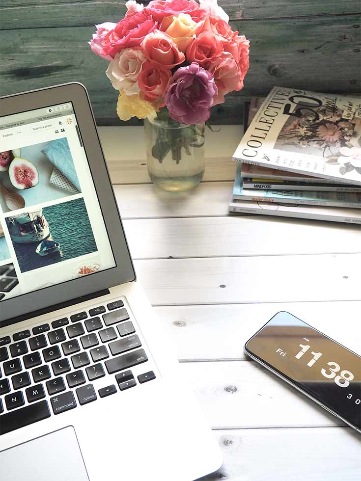 A tabletop with a laptop, cell phone, some roses and a stack of magazines.